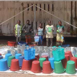 Five people are standing in front of a wooden wall, with buckets lined up in front of them 
