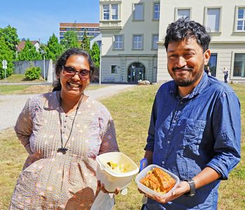 Shuba (links) und Rituparno (rechts) stehen auf einer Wiese vor dem Helmholtz-Institut Würzburg. Im Hintergrund kann man ein Teil des Gebäudes, sowie einen klaren blauen Himmel sehen. Sie lächeln beide in die Kamera mit jeweils einer Tupperbox voller Essen in der Hand.