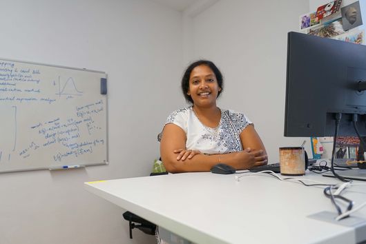 Shuba standing behind a white desk, with her arms relaxing on it in a crossed manner. She is smiling into the camera. On the desk, you can see her computer setup and a big coffee mug. A whiteboard with formulas, graphs and other notes is hanging at the back of the room. The wall next to her desk is decorated with a colorful variety of post cards.