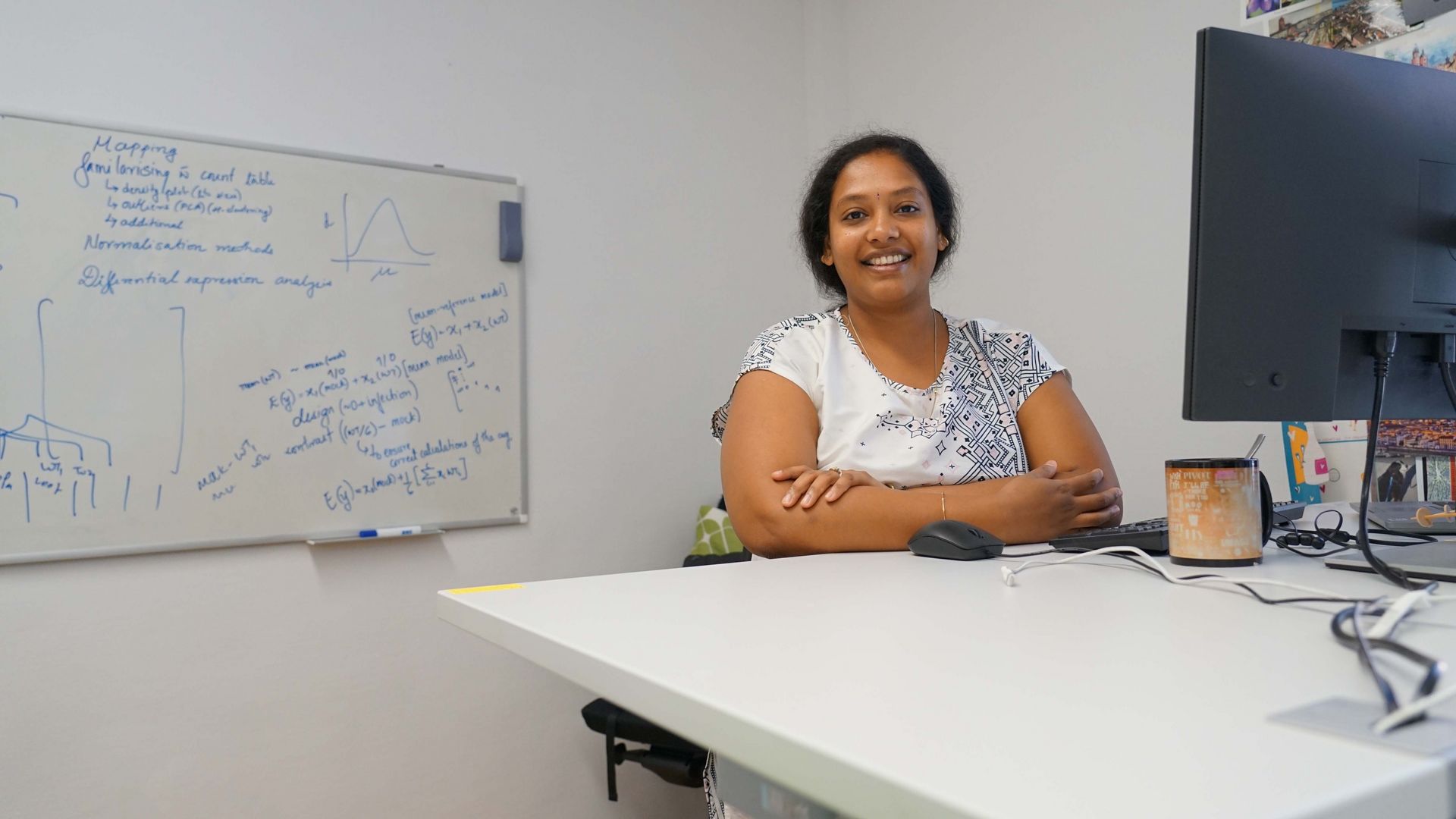 Shuba standing behind a white desk, with her arms relaxing on it in a crossed manner. She is smiling into the camera. On the desk, you can see her computer setup and a big coffee mug. A whiteboard with formulas, graphs and other notes is hanging at the back of the room. The wall next to her desk is decorated with a colorful variety of post cards.